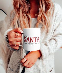 a woman holding a white coffee mug with the words dear santa on it in red