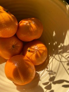 four oranges sitting in a white bowl on a table with shadow from the sun