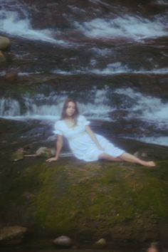 a woman sitting on top of a mossy rock next to a river filled with water