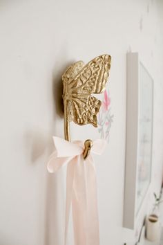 a gold butterfly on a white wall with pink ribbon hanging from it's back