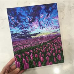 a hand holding up a card with pink flowers in the background and purple clouds above it