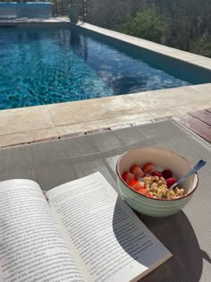 a bowl of fruit and cereal next to an open book on a table near a swimming pool