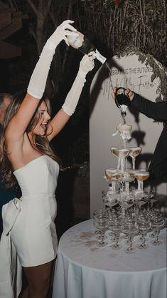 a woman in white dress standing next to a table with wine glasses on top of it