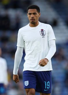 a man in white shirt and blue shorts standing on soccer field with his hands behind his back