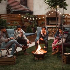 group of people sitting around a fire pit in the backyard with string lights strung over it