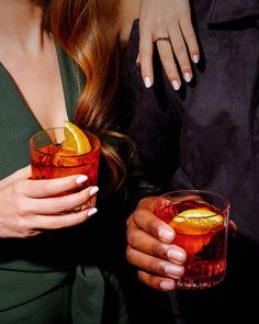 two women holding glasses filled with drinks and orange slices on the rims, one woman's hand resting on her shoulder