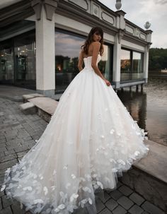 a woman in a white wedding dress standing on the sidewalk near water with her back to the camera