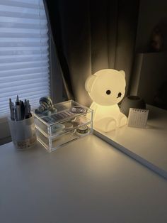 a white teddy bear sitting on top of a desk next to a box with pens and pencils