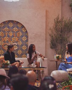 three women sitting on chairs talking to each other in front of a group of people