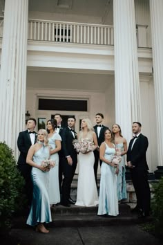 a group of people standing in front of a white building wearing formal wear and tuxedos