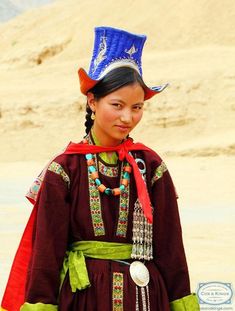 a young woman in traditional dress and headdress stands on the sand dunes, looking at the camera