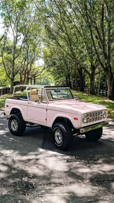 A classic Ford Bronco in a unique Rosé pink color, parked on a shaded road lined with lush green trees. The Bronco features a white soft top, large off-road tires, and a clean, well-maintained body, showcasing its retro charm and stylish color palette. Classic Car Convertible, Pink Ford Truck, Cute First Cars, Dream Car Jeep, Vintage Ford Bronco, Old Bronco, Bow Art, Dream Cars Jeep, Car Goals