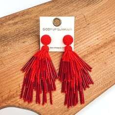 Circle beaded stud earrings with a beaded tassel in red. These earrings are pictured laying on a brown piece of wood on a white background. Sale Items, Night Out