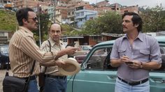 two men standing next to each other in front of a green car with another man holding a hat