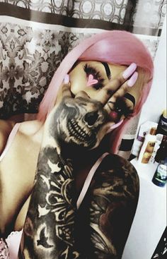 a woman with pink hair and tattoos on her body sitting in a chair holding her hand up to her face