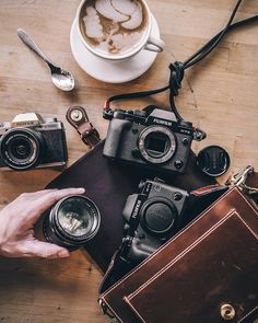 a person holding two cameras next to a cup of coffee and other items on a table