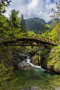 a wooden bridge over a river surrounded by trees and mountains in the distance with water running under it