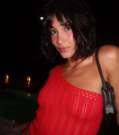 a woman in a red top standing next to a pool at night with her hand on her hip