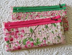 three pink and green zippered purses sitting on top of a white quilt covered bed