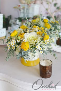 yellow and white flowers are arranged in a vase on a table with a candle next to it