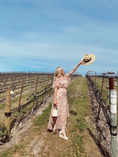 a woman in a dress and hat standing on a dirt road next to rows of vines