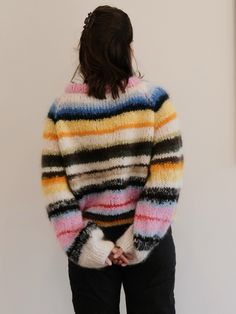 a woman standing in front of a white wall wearing a colorful sweater and black pants