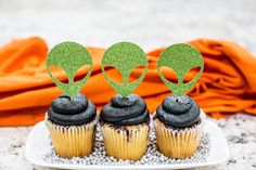 three cupcakes with black frosting and green decorations on top, sitting on a plate