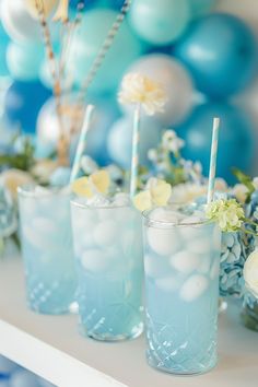 three glasses filled with blue liquid sitting on top of a table next to balloons and flowers