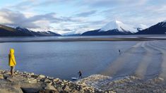 two people in yellow jackets are standing on rocks near the water with mountains in the background