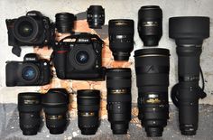 many different types of cameras and lenses lined up against a brick wall, all with their hoods open