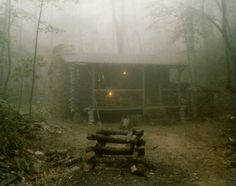 a cabin in the woods on a foggy day with some logs stacked around it