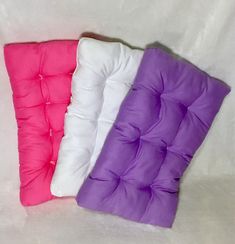 three different colored pillows sitting next to each other on a white surface with one pink, one purple and one white