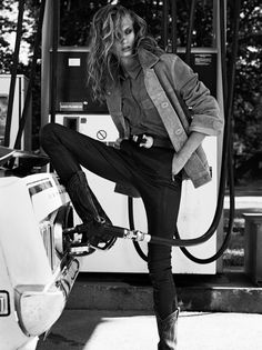 a woman in black pants and jacket leaning on a gas pump with her legs crossed