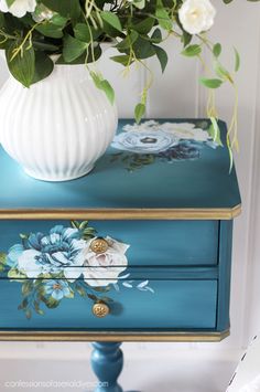 a white vase with flowers sitting on top of a blue painted dresser next to a green plant