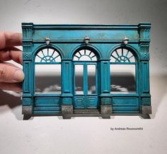 a hand holding up a miniature building made out of plastic and wood with arched windows