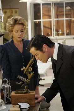 a man looking through a microscope while standing next to a woman in a black dress