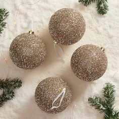 three glittered christmas ornaments with white ribbon on snow covered ground next to evergreen branches