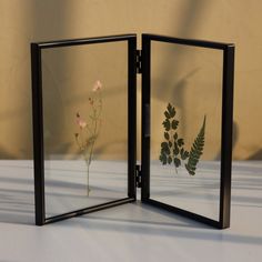 two black frames with flowers and plants in them on a white tableclothed surface