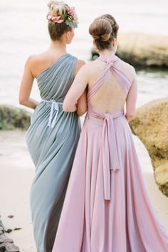 two women in dresses standing next to each other on the beach looking at the water