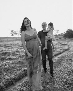 a pregnant woman in a long dress is walking with her husband and baby daughter through a field