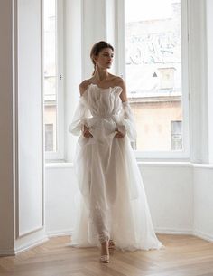 a woman standing in front of a window wearing a white dress with ruffled sleeves