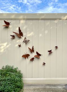 several metal butterflies are hanging on the side of a white fence with a green plant in front of it