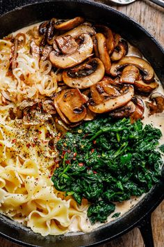 a pan filled with pasta, mushrooms and spinach on top of a wooden table