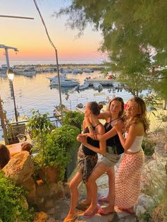 three young women are standing on the steps by the water at sunset or sunrise, with boats in the background