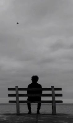 a person sitting on a bench looking out at the ocean with a kite flying in the sky