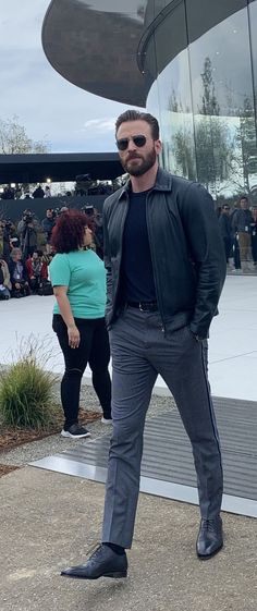 a man standing in front of a building wearing sunglasses and a black jacket with his hands in his pockets