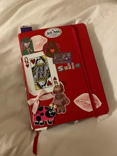 a red notebook covered in stickers and paper on top of a white bed sheet