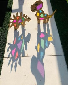 two paper cut out dinosaurs on the ground with long shadows from their tails and legs