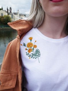 a woman wearing an orange jacket and white shirt with flowers on the front is standing near water