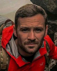 a man wearing a red jacket and backpack standing in front of some rocks with his hands on his hips